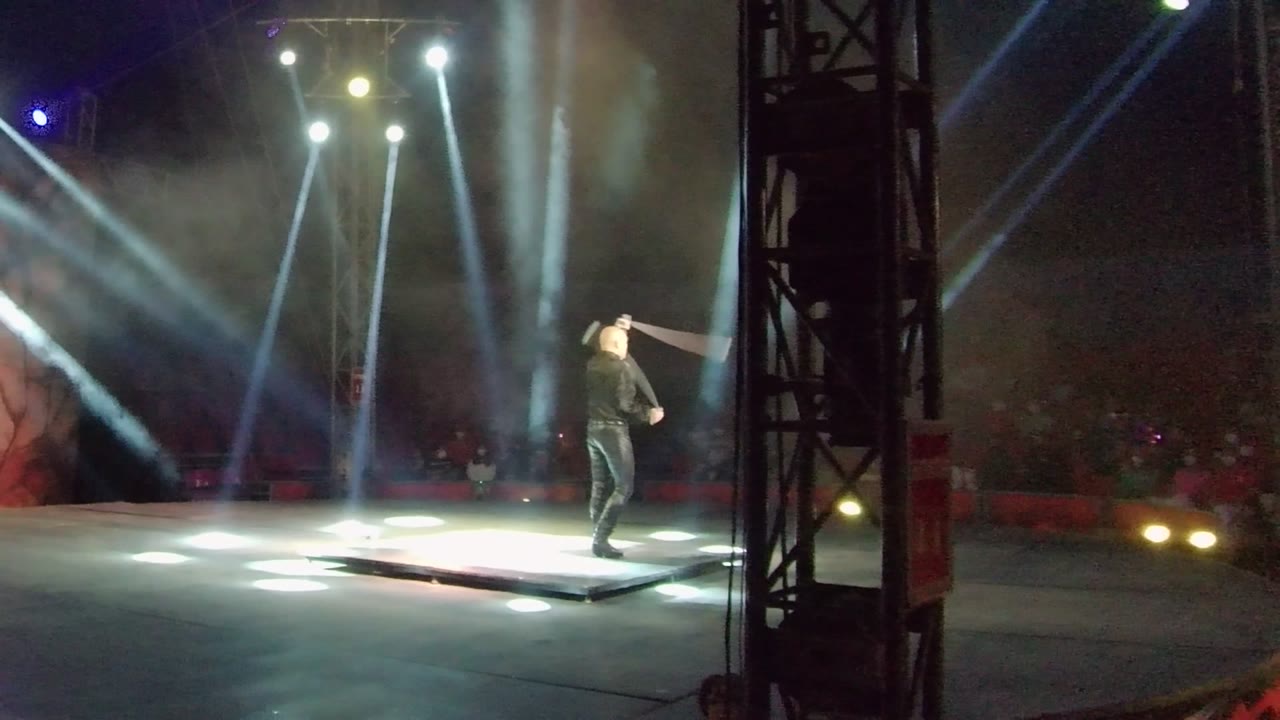 In San Jose Del Cabo, the performer turns with the large prop held across the body while the illuminated stage platform and overhead beams dominate the frame.