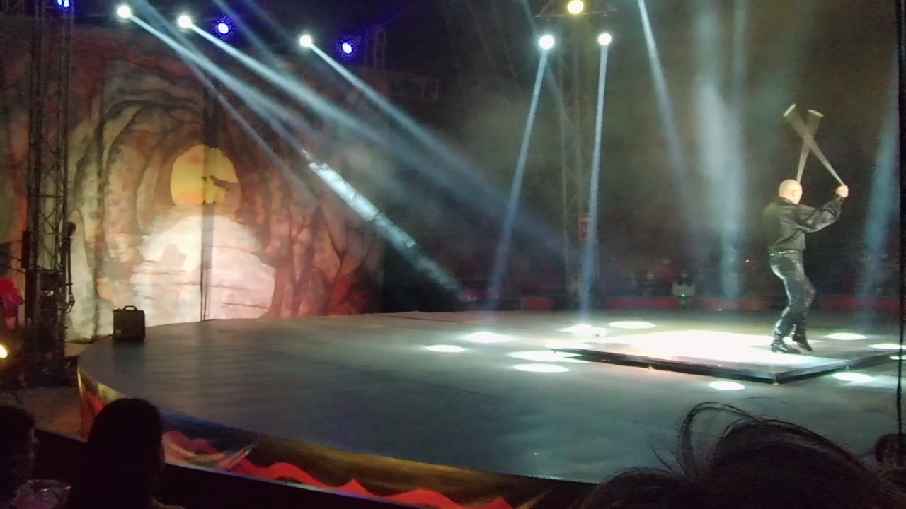 In San Jose Del Cabo, Mexico, a black-clad stage performer lifts a large sword- or axe-like prop while white and blue beams cross over a cave-themed backdrop.