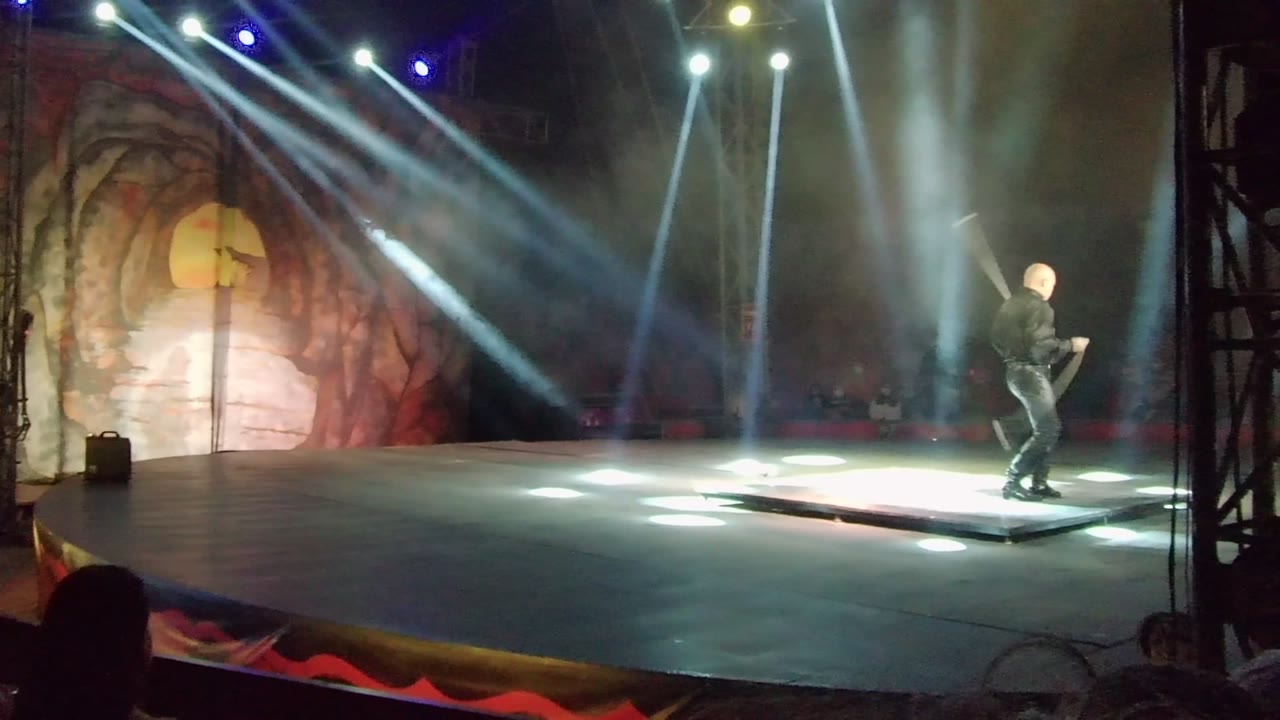 Performer swings a large prop on a wide stage with bright beams.