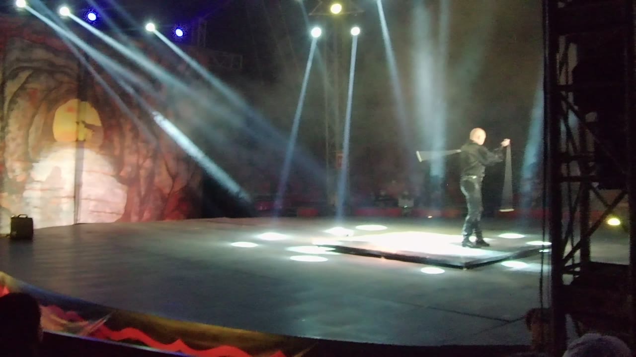 In San Jose Del Cabo, Mexico, the performer appears near the right side of the lit platform with the prop lifted as stage beams fan across the dark seating area.