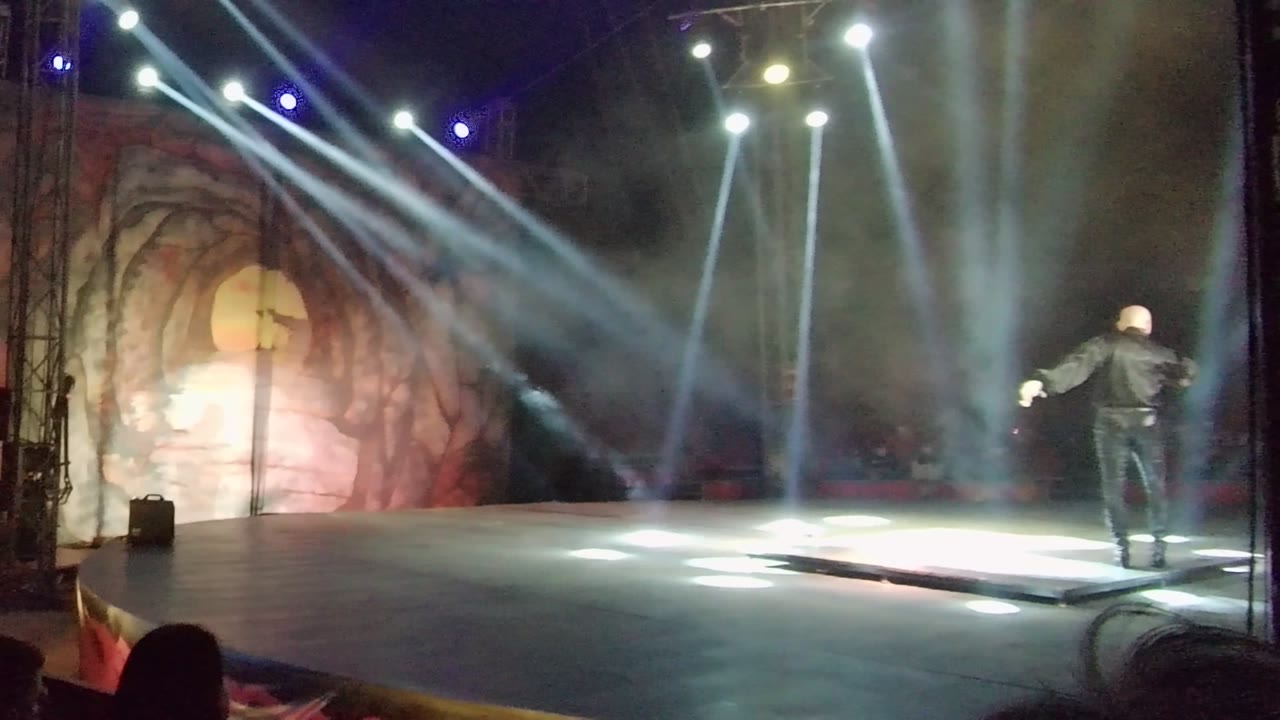 A black-clad performer stands near center stage in San Jose del Cabo while white beams cross through haze above a painted backdrop and audience silhouettes line the foreground.