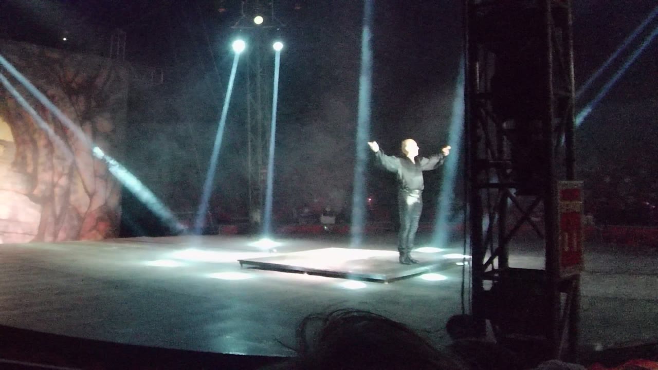 A solo performer in dark clothing stands with arms outstretched on a San Jose Del Cabo stage as white beams shine through haze from above.