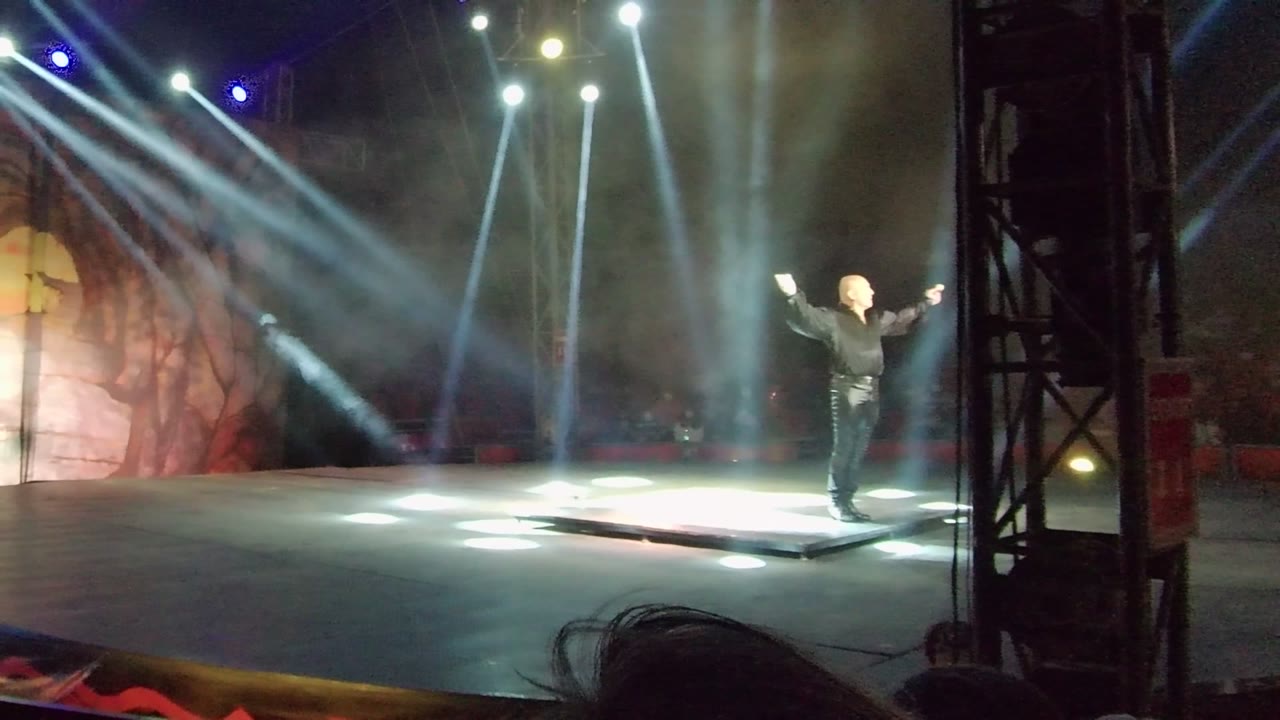 A wide stage view in San Jose Del Cabo shows the performer slightly right of center beside a bright floor-lit area and vertical rigging at frame right.