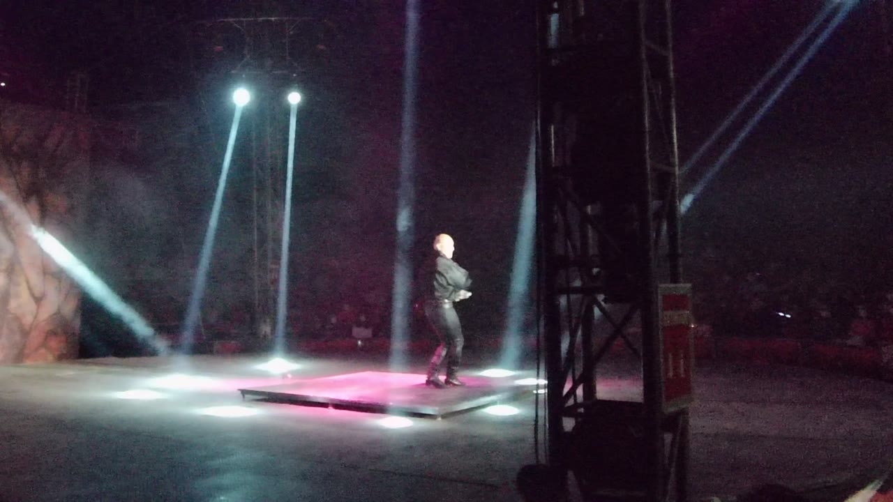 The performer moves across the San Jose del Cabo stage as blue-white beams cut through haze above a pink-lit platform.