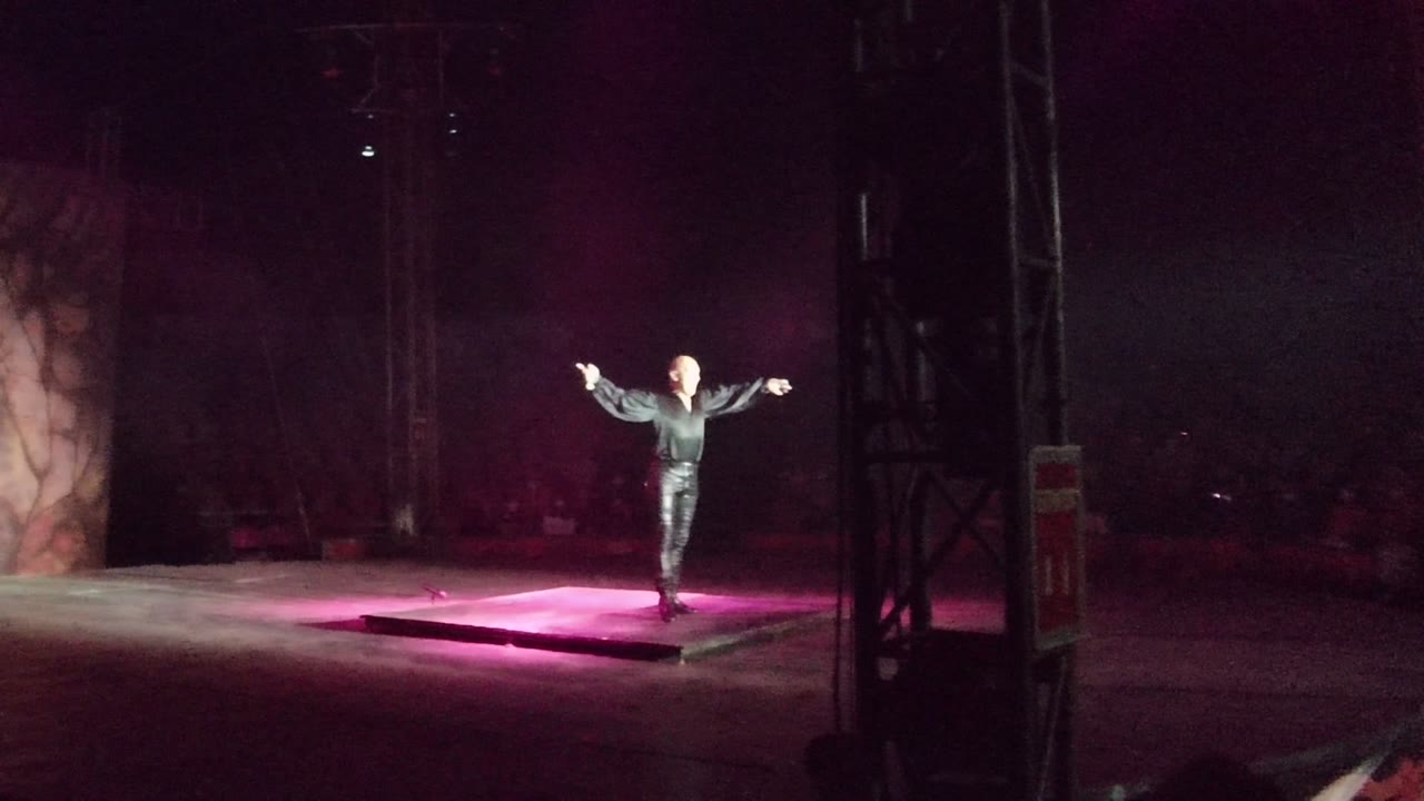 A performer stands on the San Jose del Cabo stage with arms extended, surrounded by pink floor light and a dark audience background.