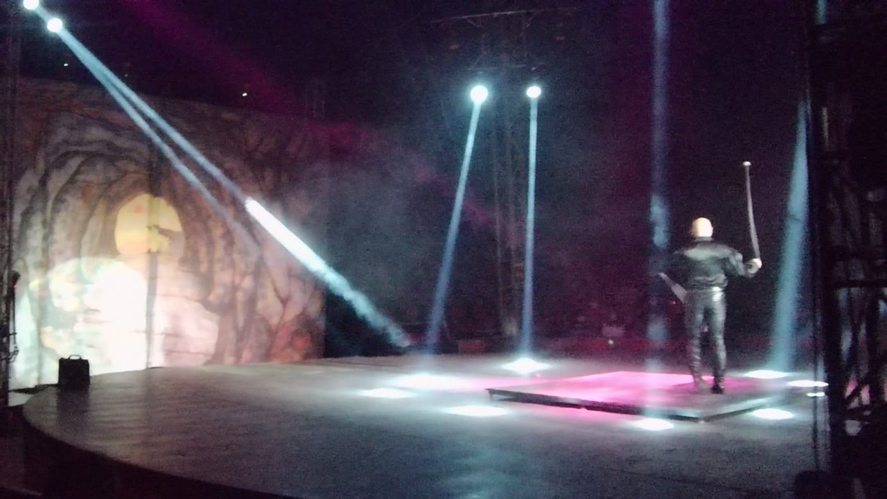 Wide view of a solo performer on a San Jose del Cabo stage, standing near bright floor lights as white beams cut through smoke against a textured backdrop.