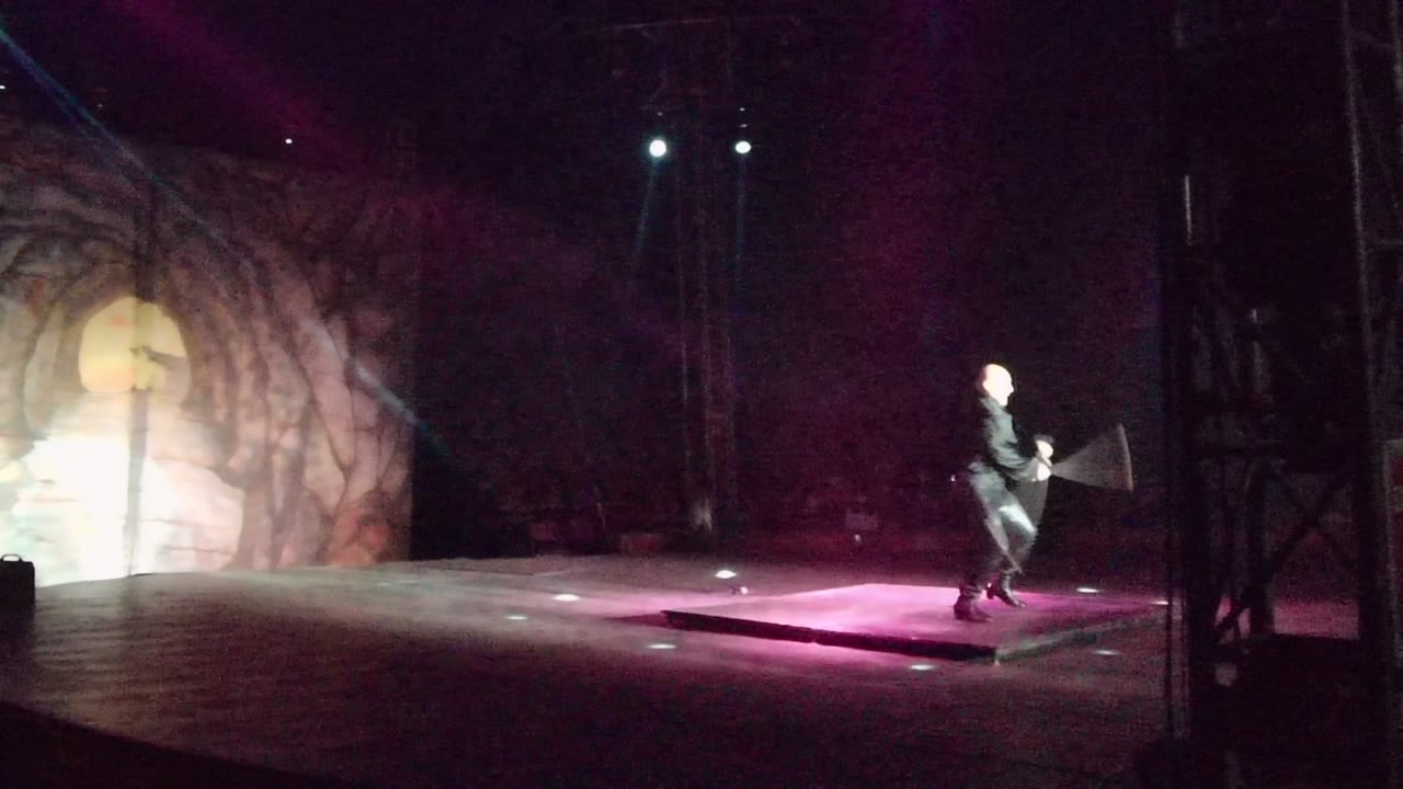 The performer reaches the right side of the San Jose del Cabo stage, with the long prop angled upward and white overhead lights shining through purple haze.