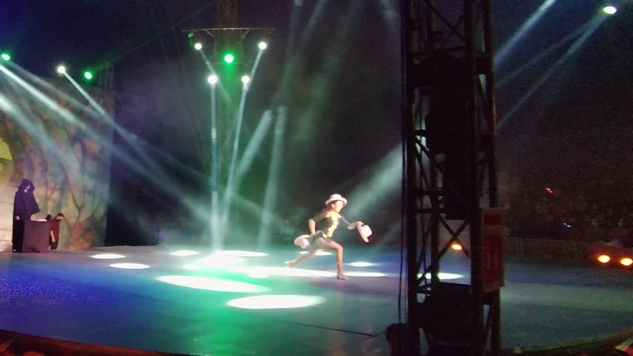 In San Jose Del Cabo, a dancer lunges low on a stage under white and green spotlights while a host stands near a podium beside a painted backdrop.