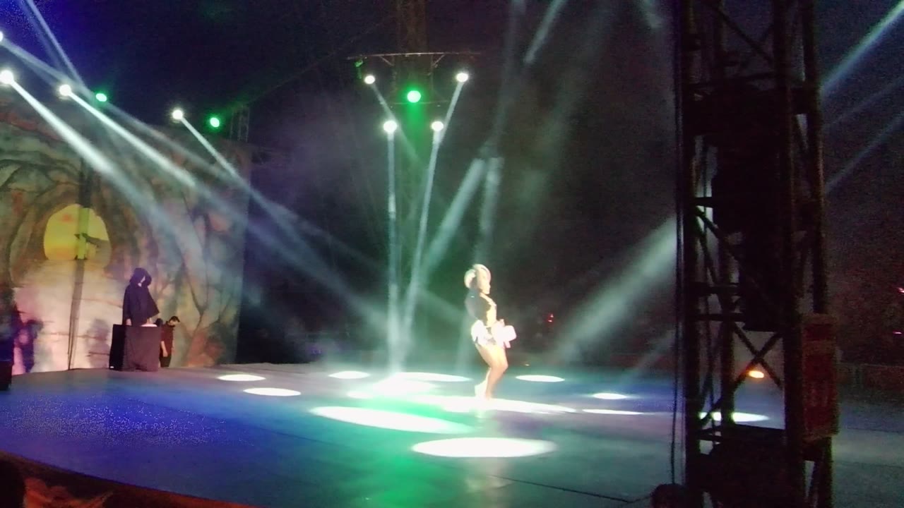 Dancer stands under bright stage beams while host remains at podium.