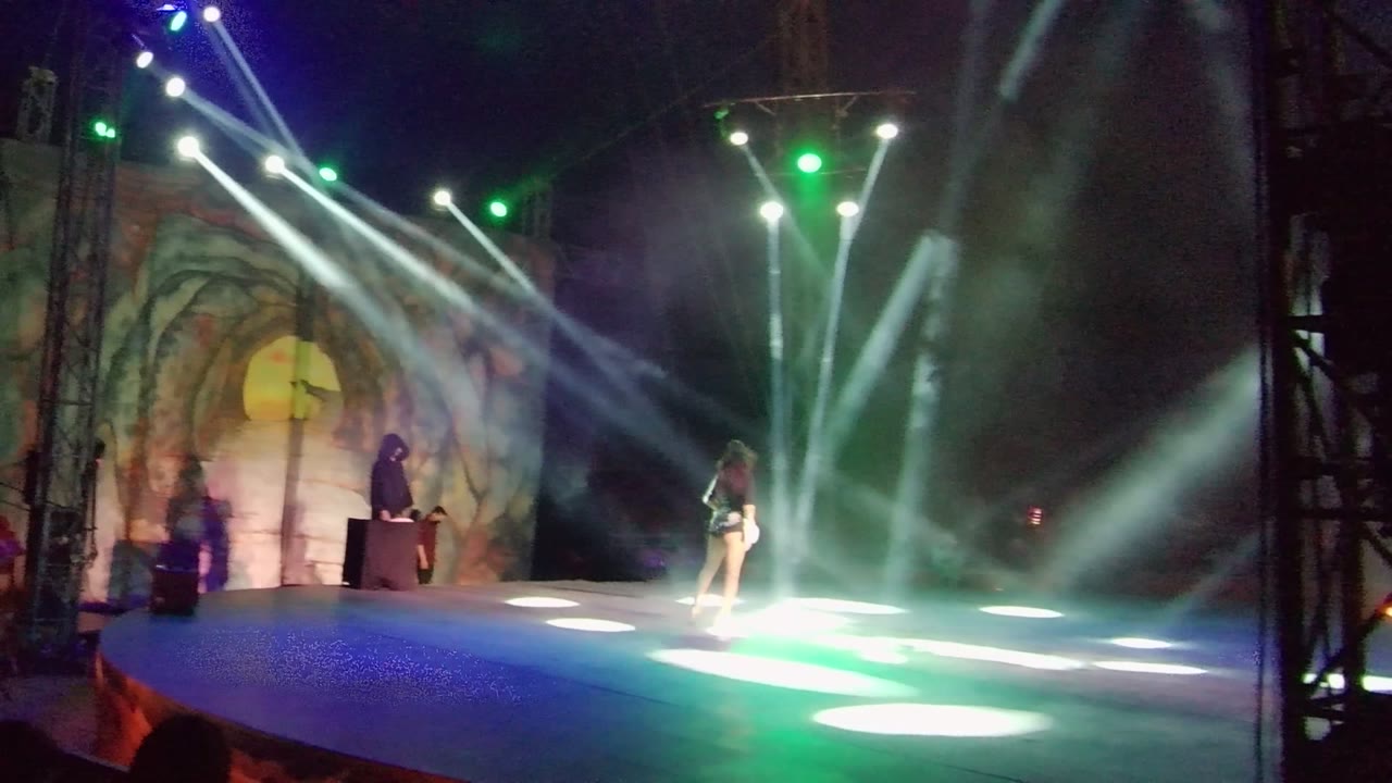In San Jose Del Cabo, the dancer moves toward the right side of the stage while multiple white beams cross overhead and the host stands near the podium at the left.