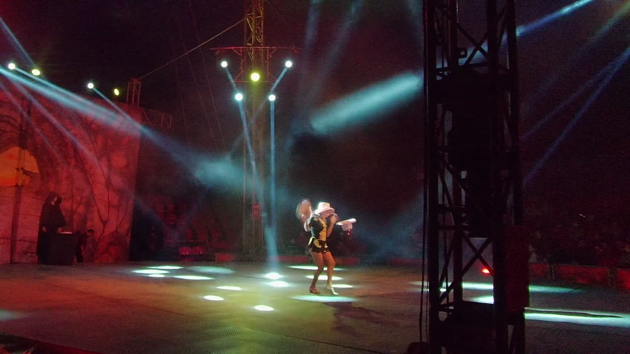 A solo performer dances on a stage in San Jose del Cabo, Mexico, with white spotlights crossing through smoke and a red scenic backdrop at left.