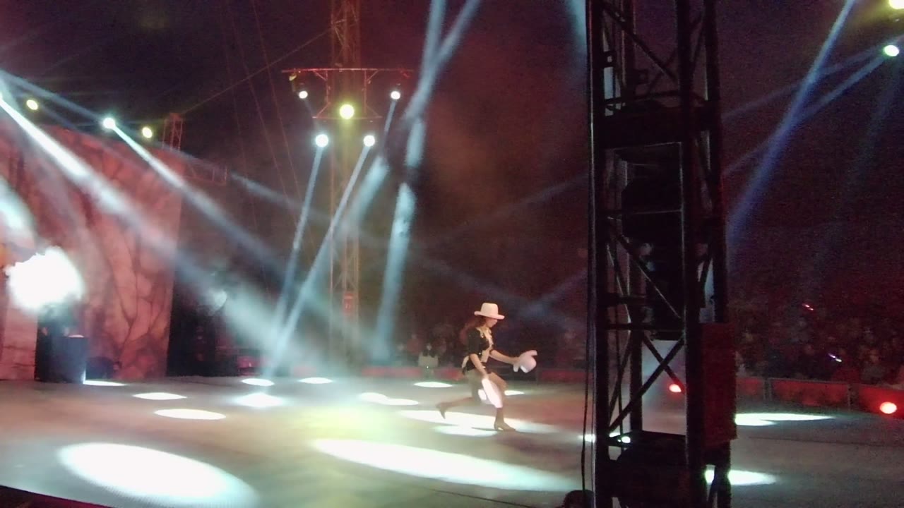Stage performer moving right under bright circular floor lights.
