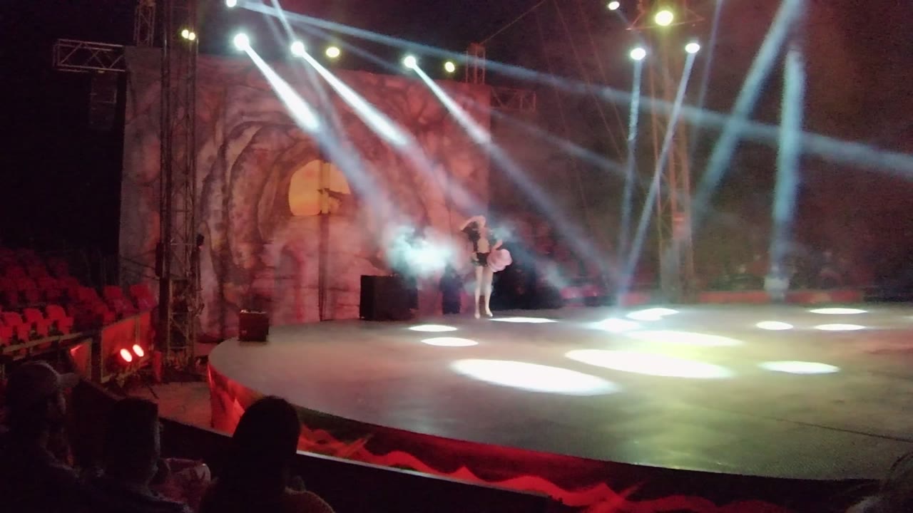Wide San Jose del Cabo stage view with a performer near the left side, red audience seats, a decorative arched backdrop, haze, and crossing white spotlights.