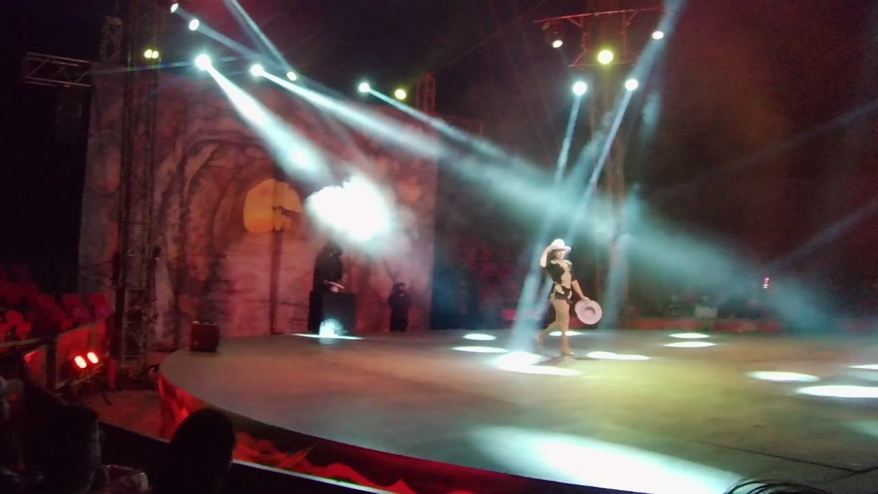 The performer stands right of center on the San Jose del Cabo stage holding a round prop as smoke and white spotlights flare near the arched backdrop.