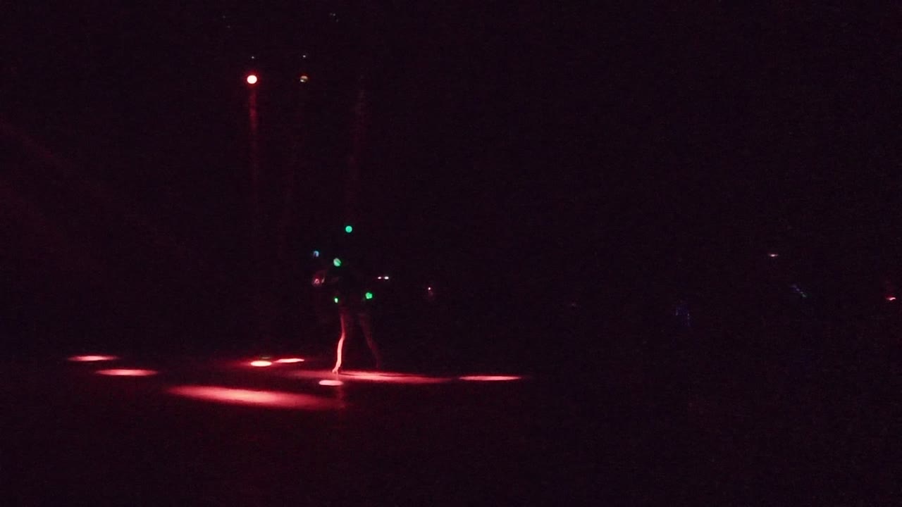 A performer with green body lights stands in a dark San Jose del Cabo stage scene as red light pools stretch across the floor.