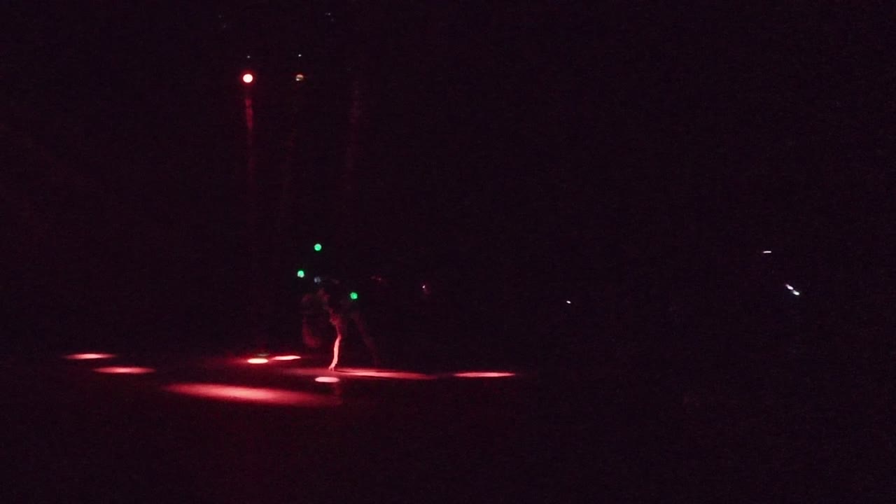The performer shifts low in the dark San Jose del Cabo stage frame, with green lights clustered near the body and red light bands on the floor.