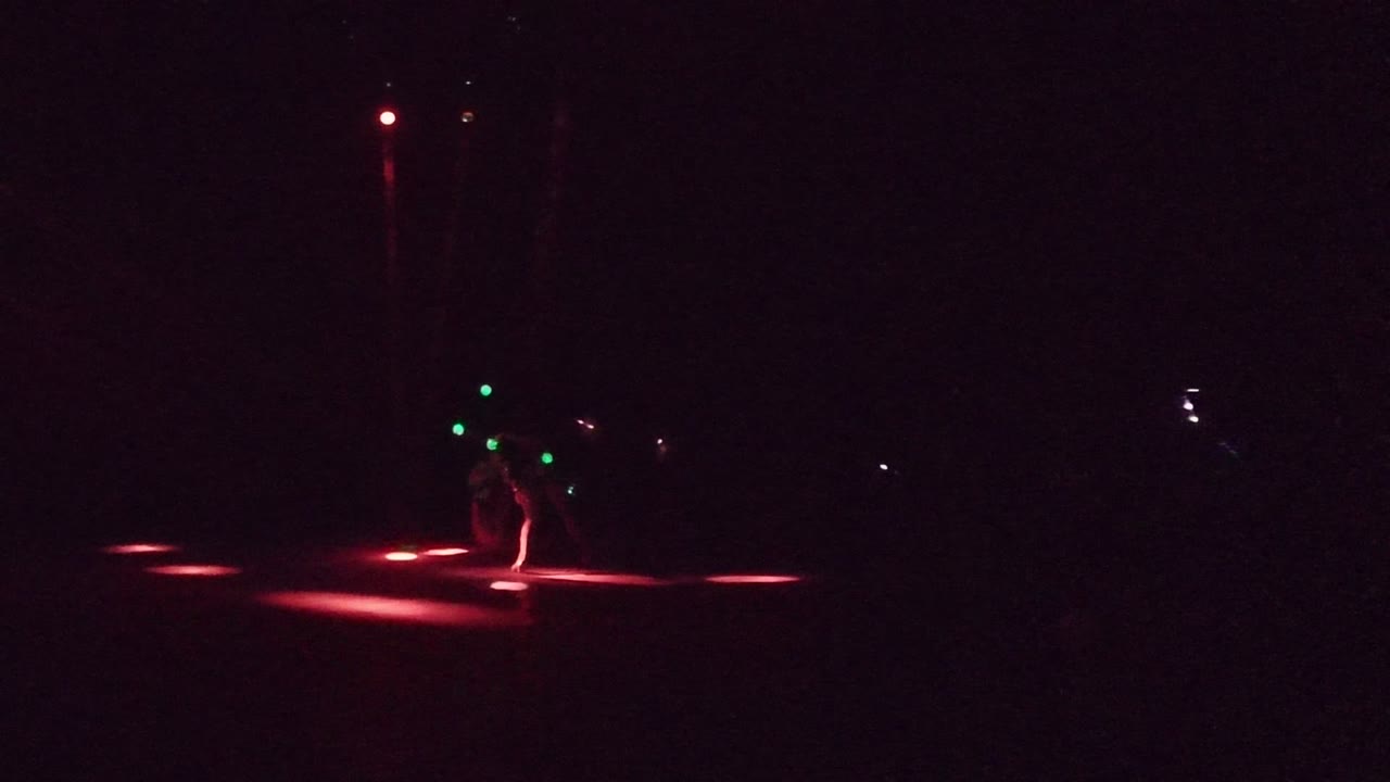 A distant dancer remains near center frame on a dark San Jose del Cabo stage while red floor spots and green body lights mark the movement.