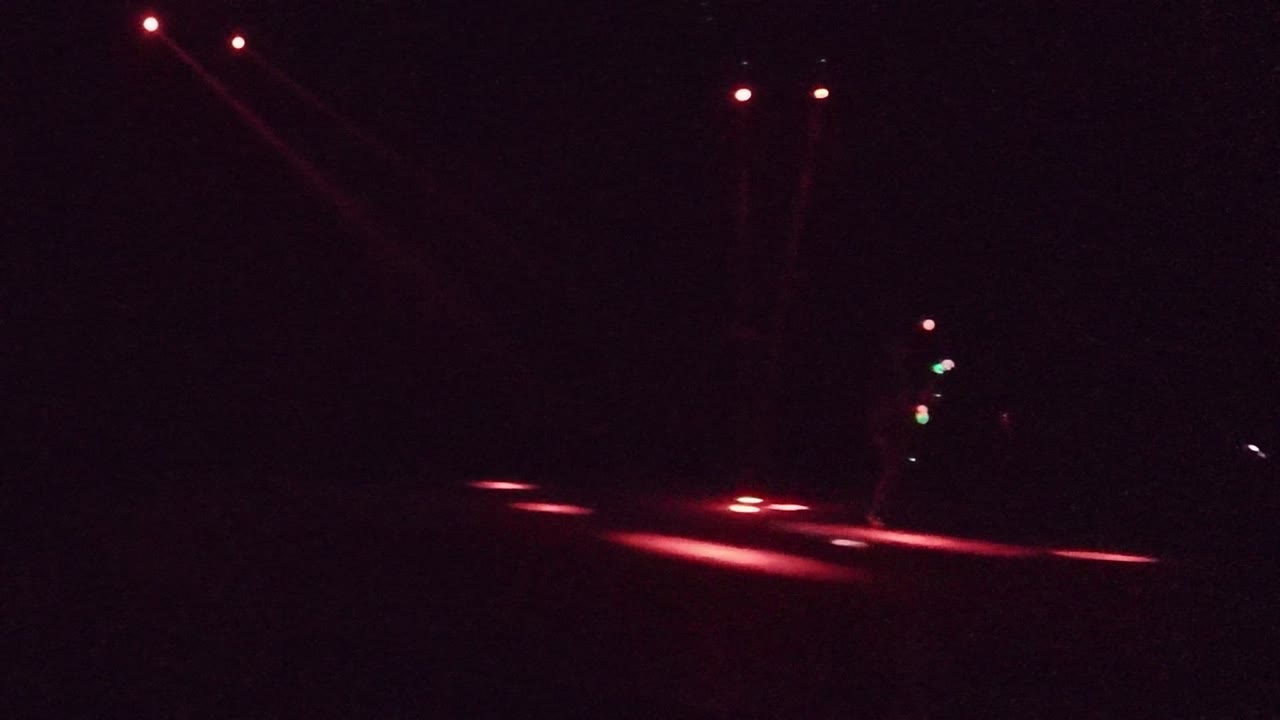 At the start of the San Jose del Cabo clip, a faint performer stands on a dark stage near red floor beams with small green and white light points visible.