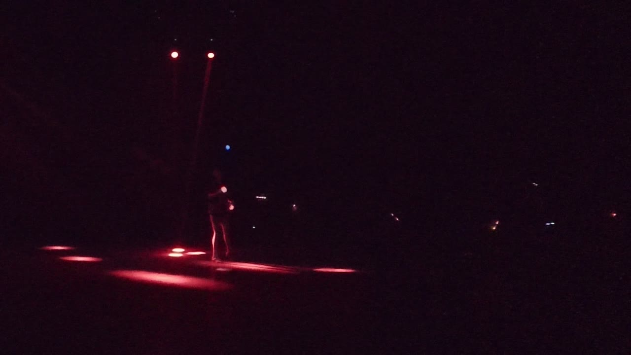 In San Jose Del Cabo, a small performer stands on a dark stage as red floor lights and faint glowing sticks mark the opening moment.