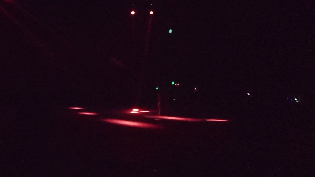 Red taillight streaks on a dark road with a faint person near a pole.