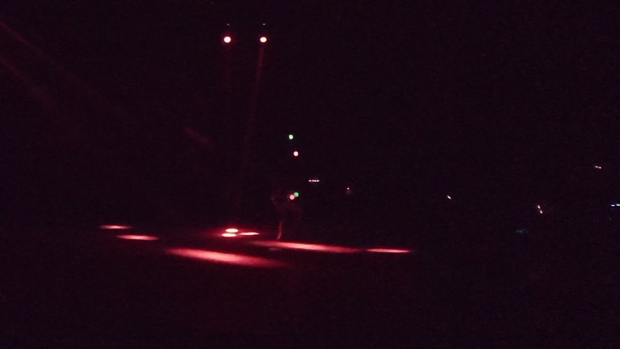 Dark San Jose del Cabo street frame with a person beside a pole, red taillight glow across the pavement, and small distant traffic lights.