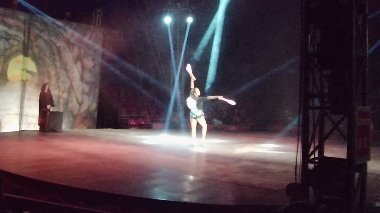 Stage performer raises a baton under crossing white light beams.