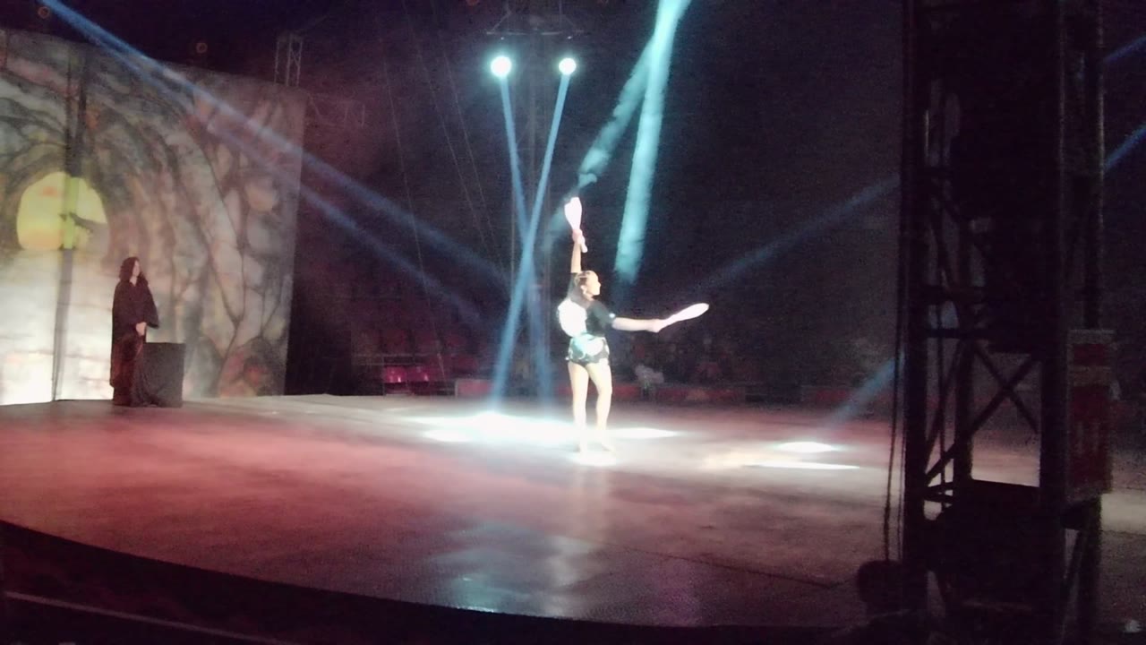 The San Jose Del Cabo stage remains brightly lit as the baton performer holds a high pose, with floor haze, a painted scenic panel, and a standing person near the podium at left.
