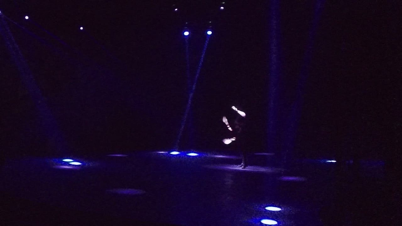 A performer in San Jose Del Cabo spins glowing staffs at the left side of a dark stage with blue floor lights and overhead beams.