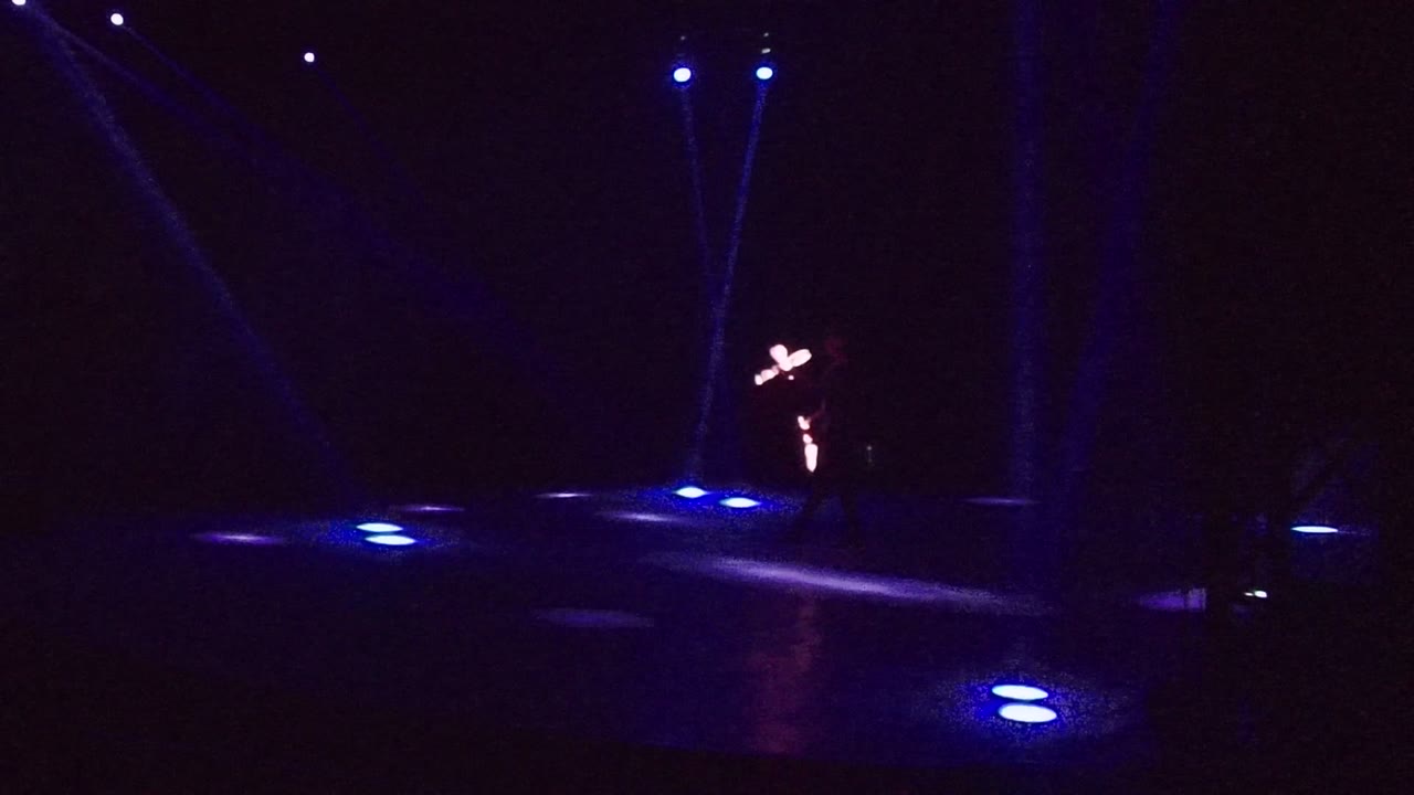 A performer stands toward the right side of the San Jose Del Cabo stage, with crossed glowing staff trails and blue light pools across the floor.