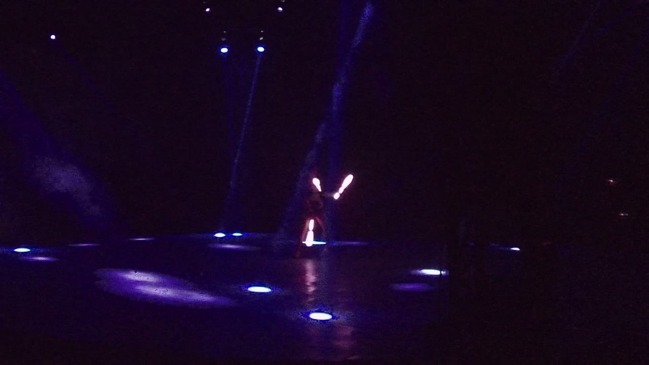 A performer in San Jose Del Cabo stands on a mostly dark stage with blue floor lights and bright glowing sticks raised near the left side of the frame.