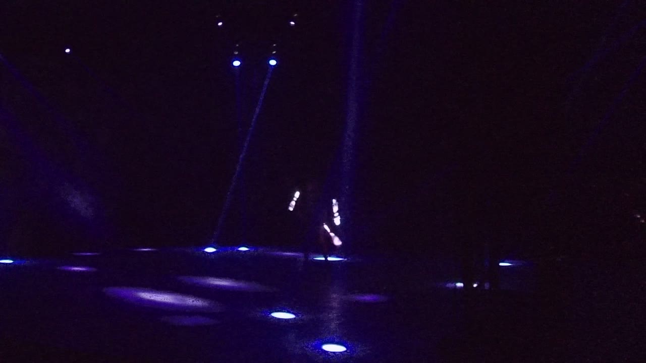 The performer in San Jose Del Cabo is small on the right side of the dark stage, with glowing sticks forming bright marks near blue floor lights.