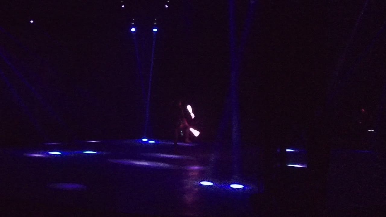 A wide dark stage in San Jose del Cabo shows blue floor lights and a small performer at left holding bright glowing sticks.