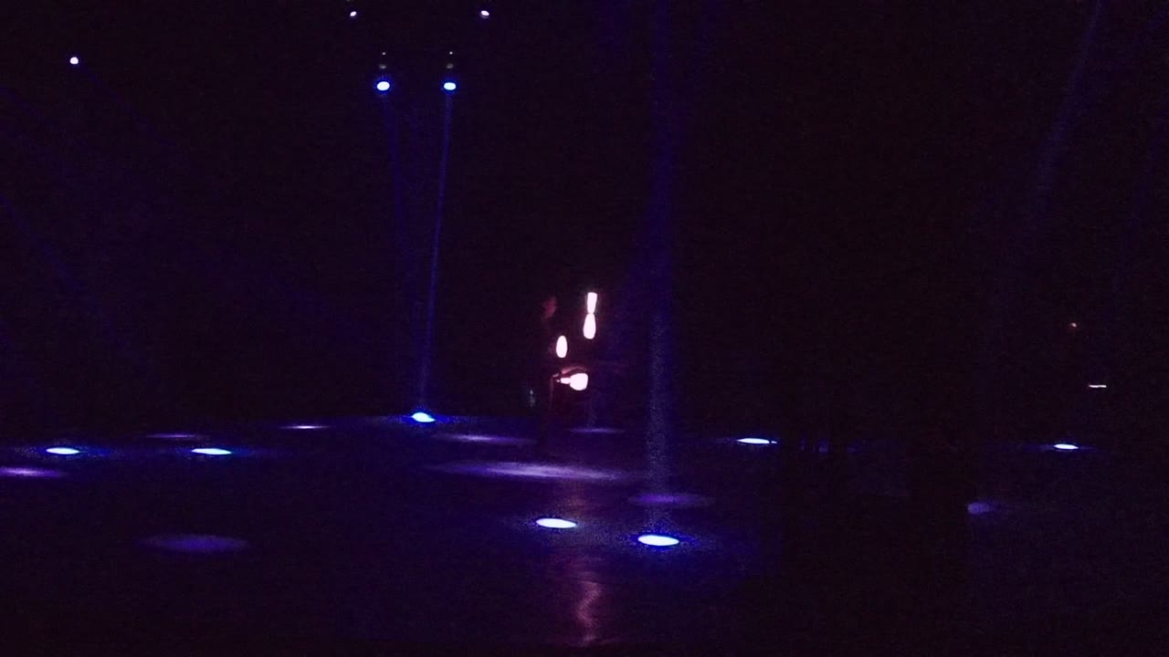 A performer on the right side of the San Jose del Cabo stage holds glowing sticks against a black background with blue floor lights across the scene.