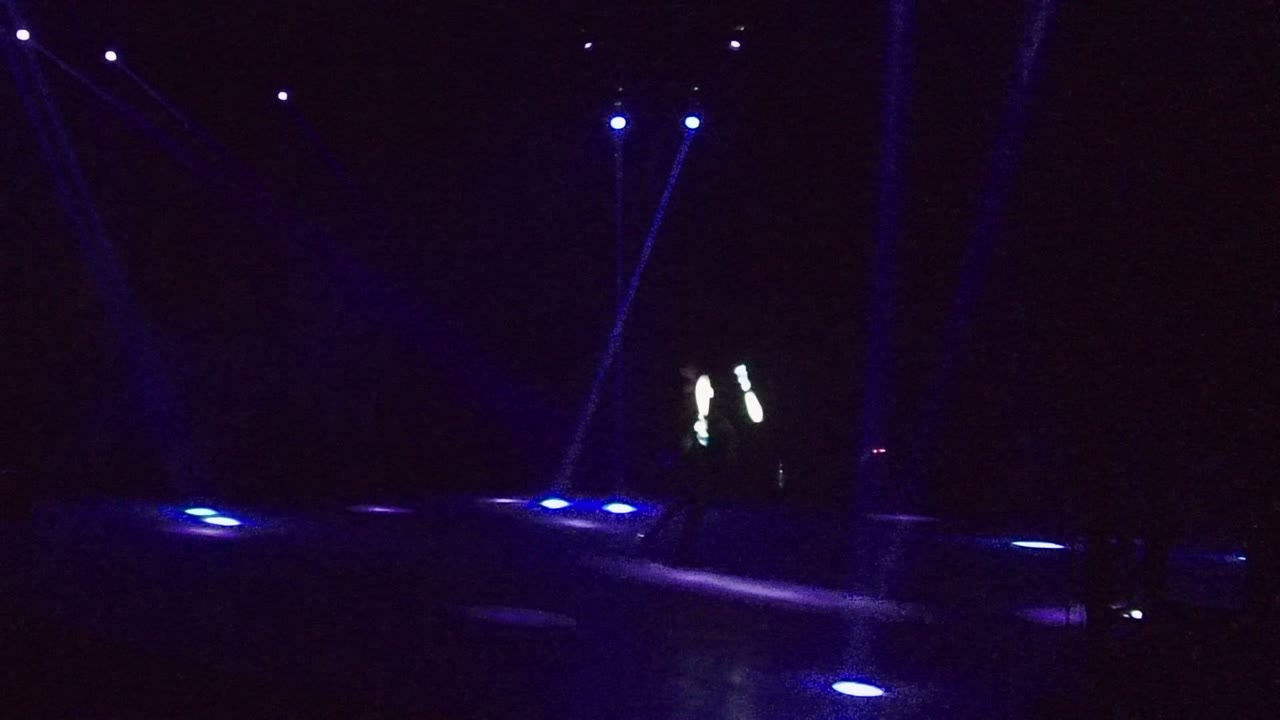 A dark San Jose del Cabo stage shows blue pools of light on the floor and a distant performer with white light wands near the left side.