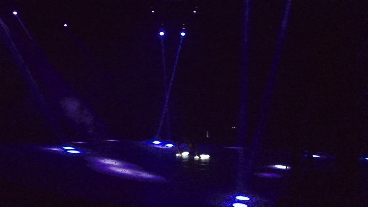 On the San Jose del Cabo stage, smoke drifts above blue floor lights while a distant performer with white light wands appears on the right.