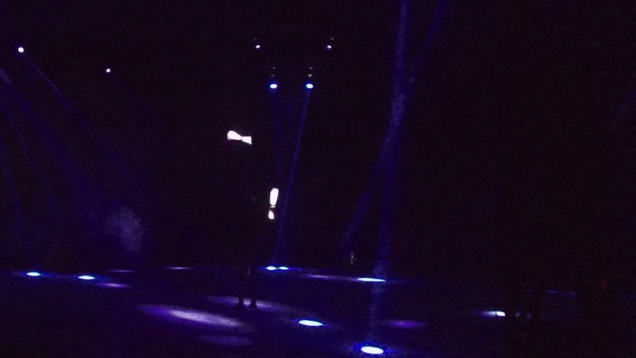 A performer in San Jose Del Cabo juggles glowing objects on a dark stage with blue floor lights and faint vertical beams.