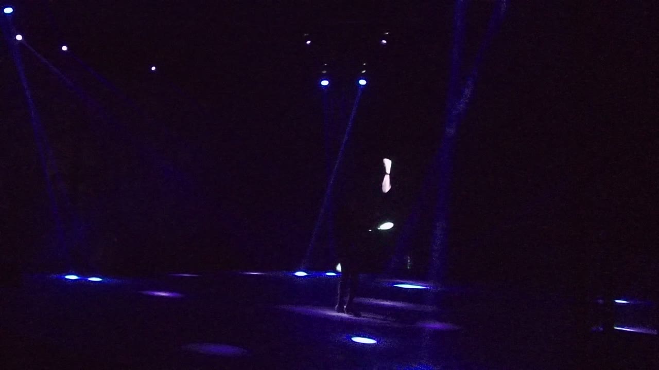 A performer stands near the right side of the San Jose Del Cabo stage while glowing white props appear against blue floor lights and a black backdrop.