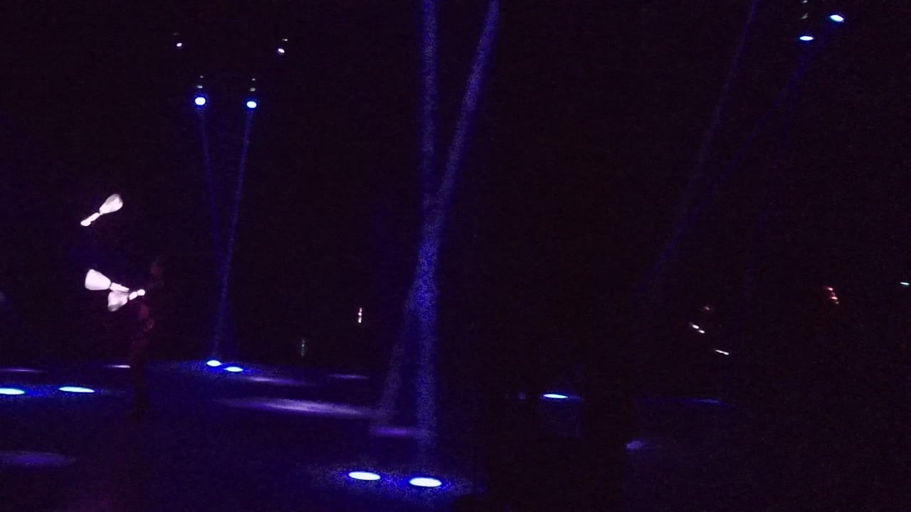 A dark San Jose del Cabo stage shows blue floor lights across the foreground and a glowing staff trail near the left side with the performer mostly hidden in shadow.