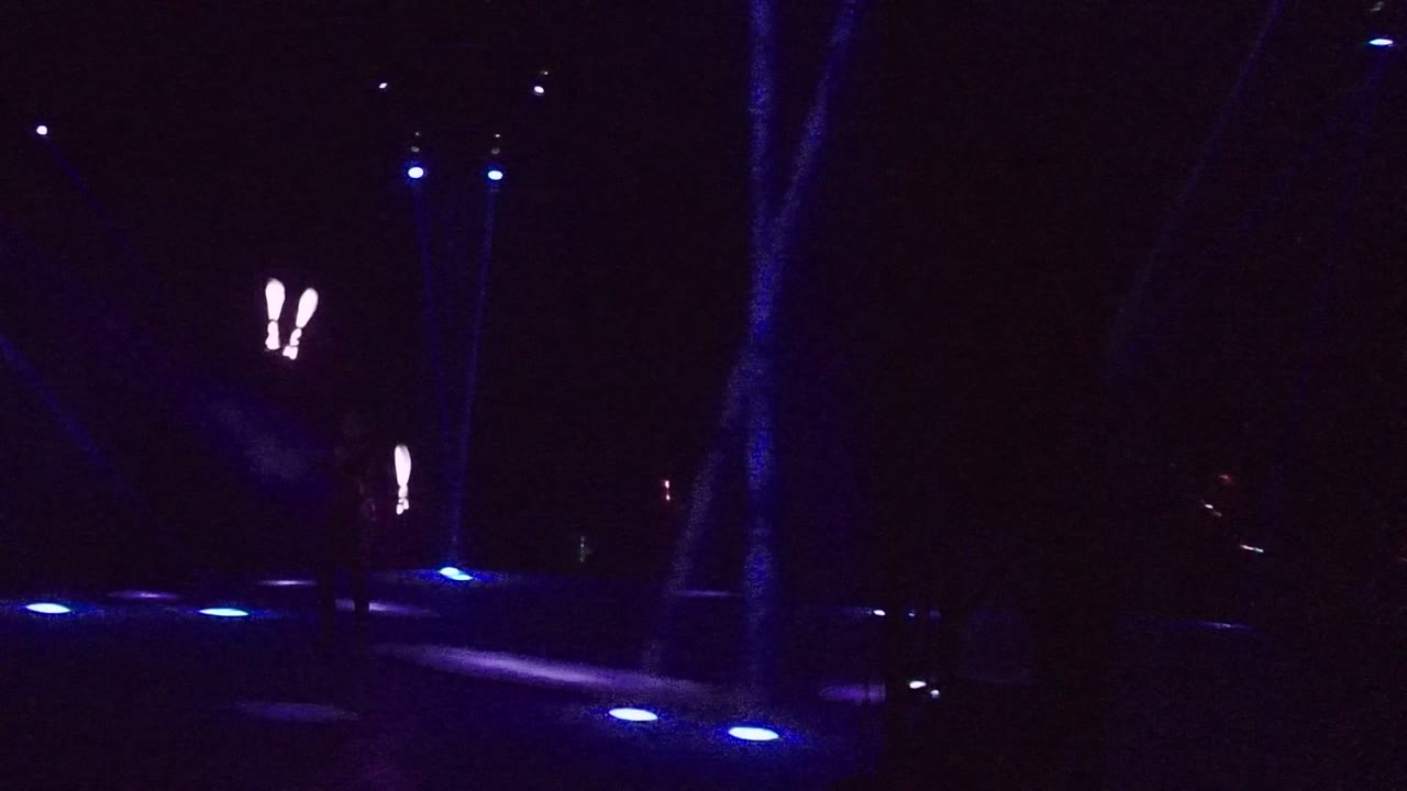 A very dark San Jose del Cabo stage shows small blue floor lights and paired bright staff marks near the center right, with the performer mostly lost in shadow.