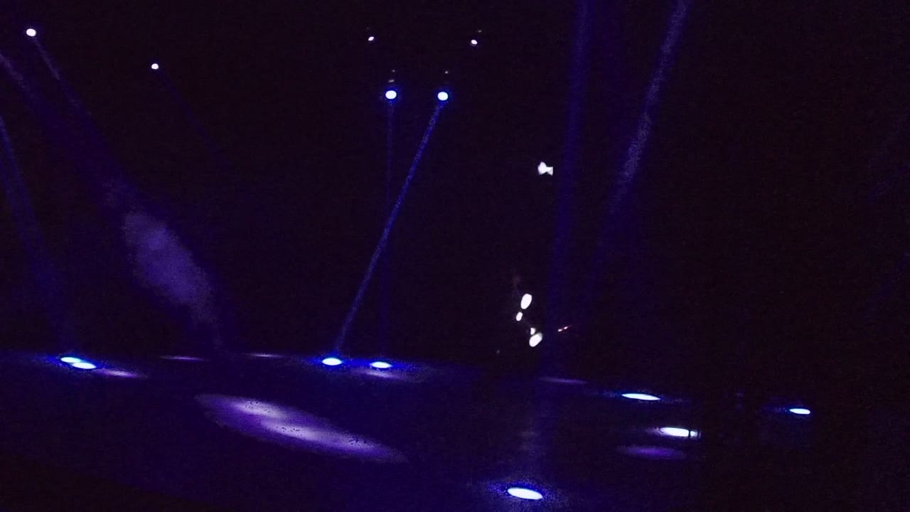 Dark stage performance in San Jose del Cabo with blue floor lights, narrow blue beams, and a mostly silhouetted performer moving white light sticks near the left side.