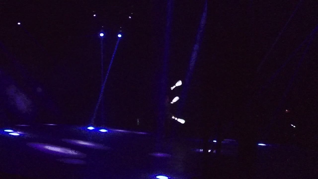 Wide dark stage in San Jose del Cabo with blue floor lights across the foreground and the performer's white light sticks moving near the right edge of frame.