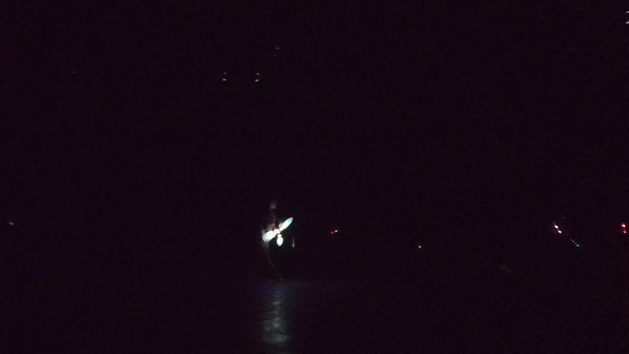 At 0:01 in San Jose Del Cabo, a small white and blue-green light cluster sits near the center over black water, with a vertical reflection beneath it and scattered distant lights.