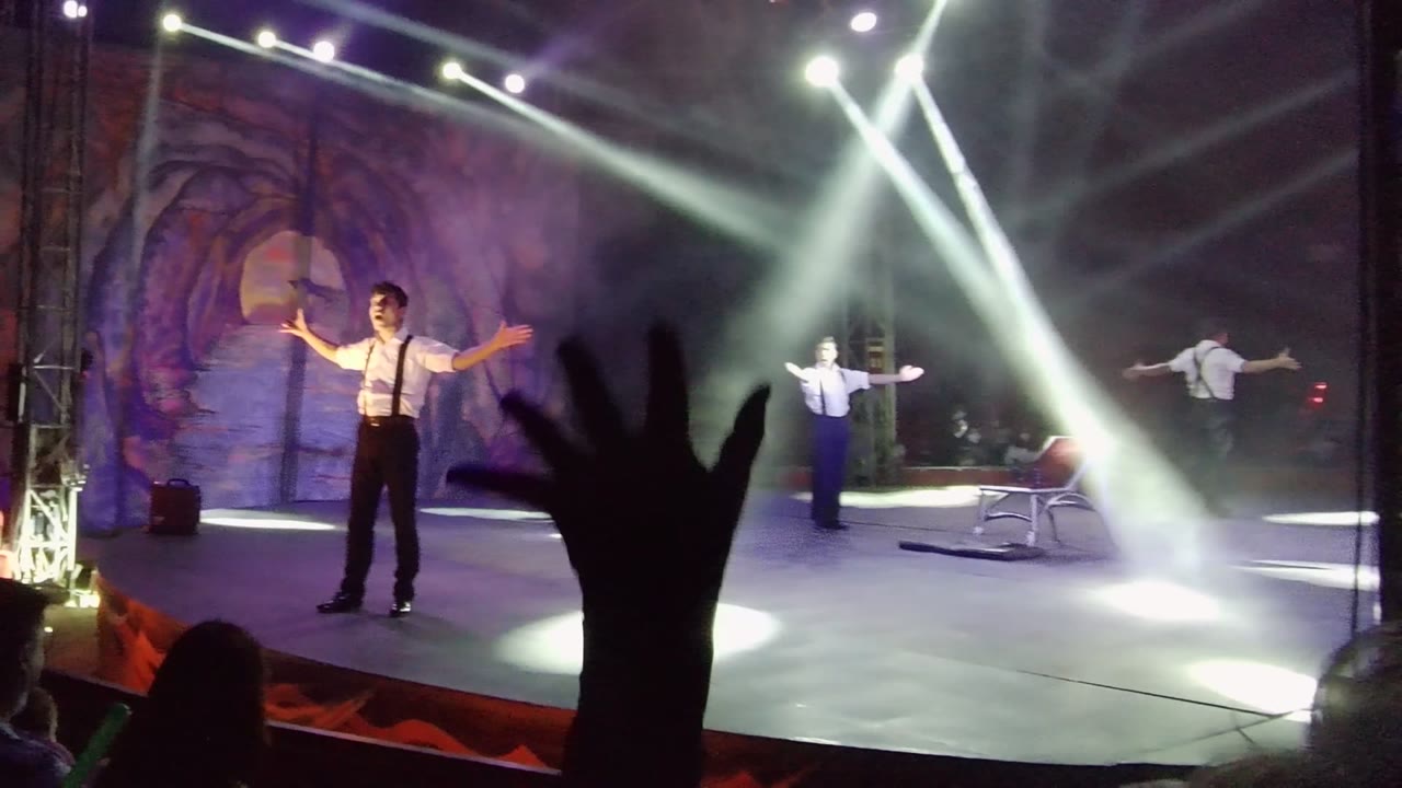 A white-shirted performer stands with arms open on a San Jose Del Cabo stage while another performer is farther back and an audience hand rises in the foreground.