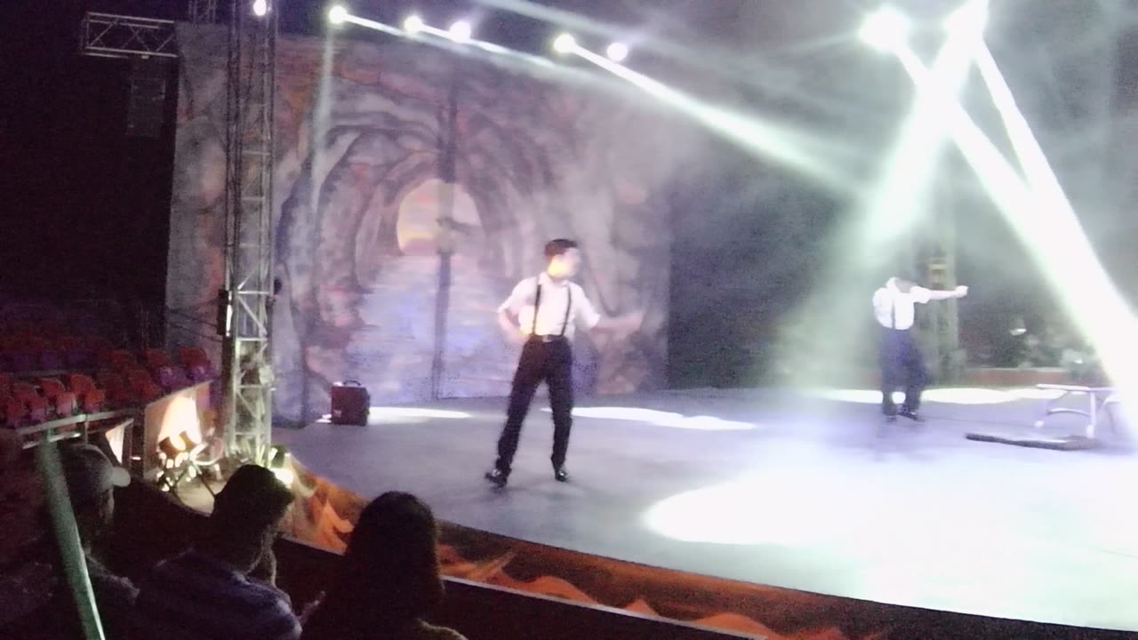 A performer moves near center stage in San Jose Del Cabo while another stands farther right and intense white beams wash out part of the stage floor.
