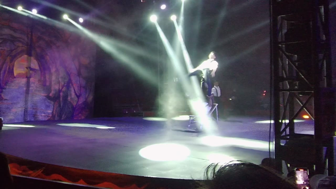 In San Jose del Cabo, Mexico, a performer balances on a chair at stage right while bright white spotlights cut through haze over a painted forest backdrop.