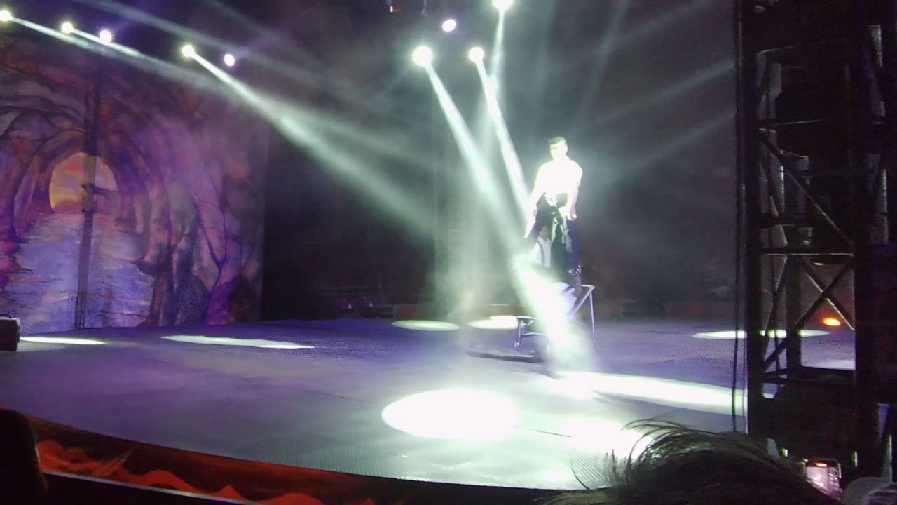 Performer standing in bright stage light beside a chair and forest backdrop.