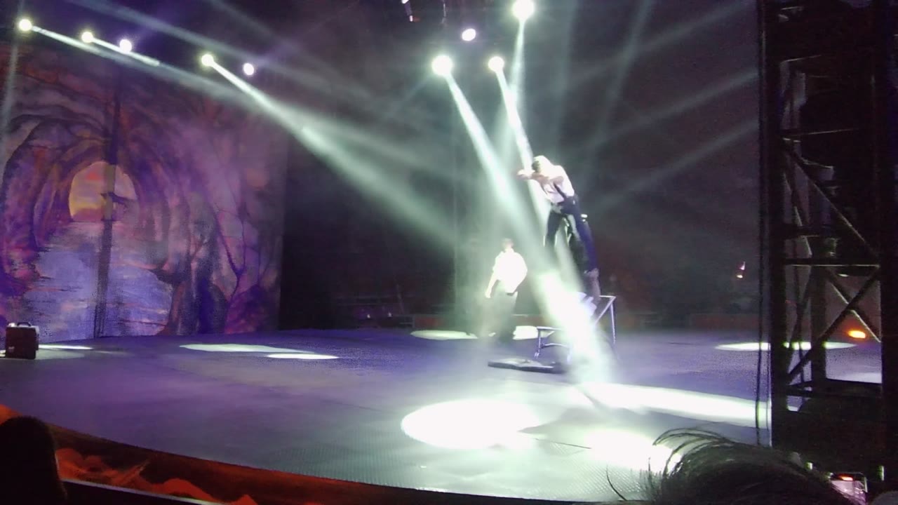 An acrobat is elevated at stage right near the chair while another performer stands farther back, with strong white beams crossing the smoky stage in San Jose del Cabo.