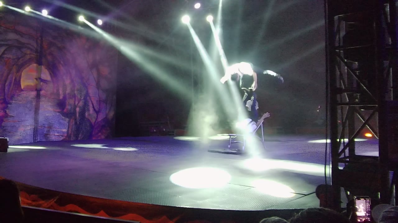 A San Jose Del Cabo stage performer leans forward during the hat trick while multiple white beams cross above the dark stage and forest backdrop.