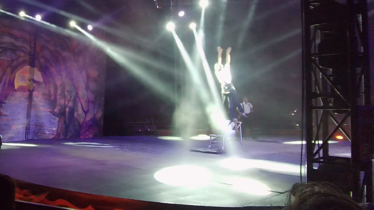In San Jose Del Cabo, Baja California Sur, Mexico, two visible performers in white hold an acrobatic lift on a stage with a forest-themed backdrop and bright beams crossing overhead.