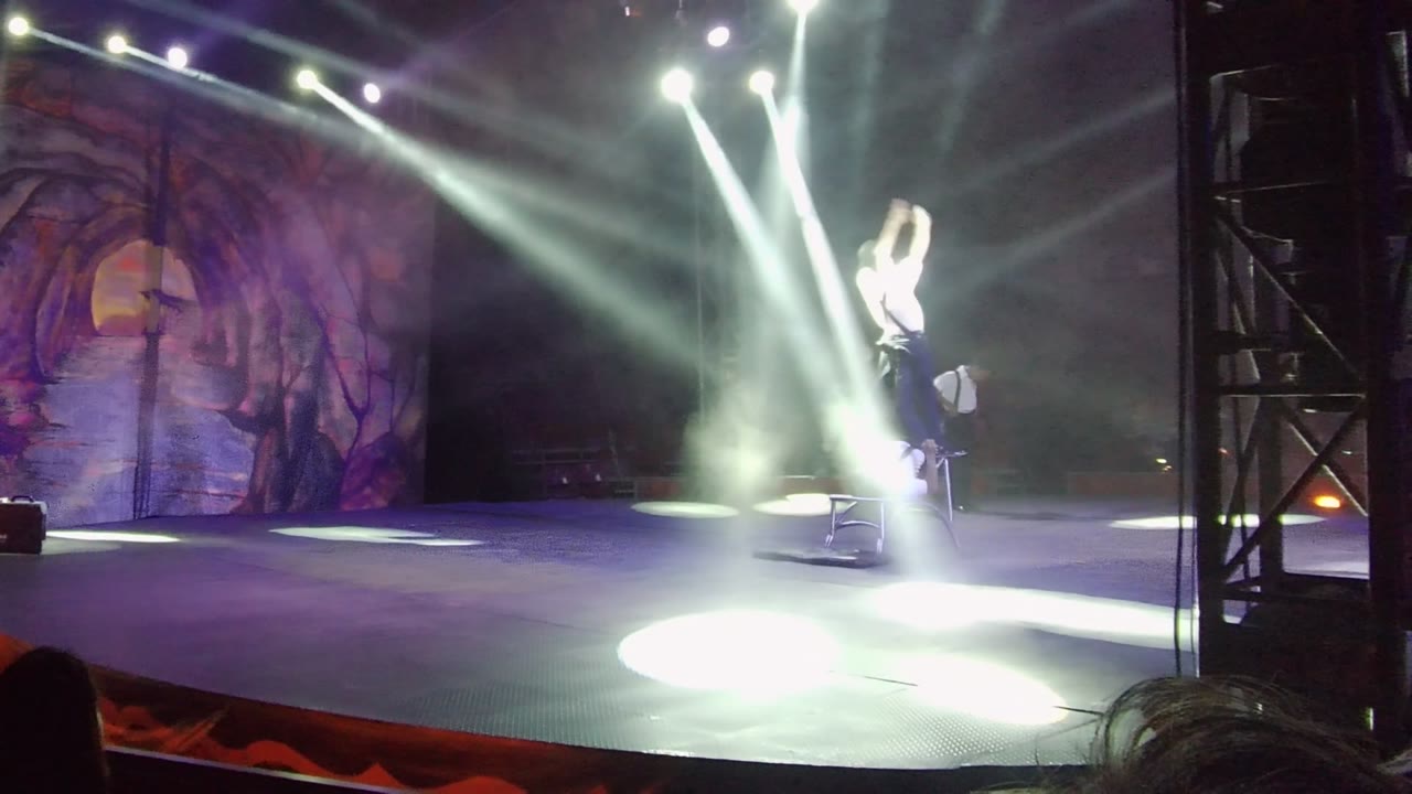 In San Jose Del Cabo, Baja California Sur, Mexico, one performer remains elevated beside the supporting performer as intense white beams flare across the right side of the stage.